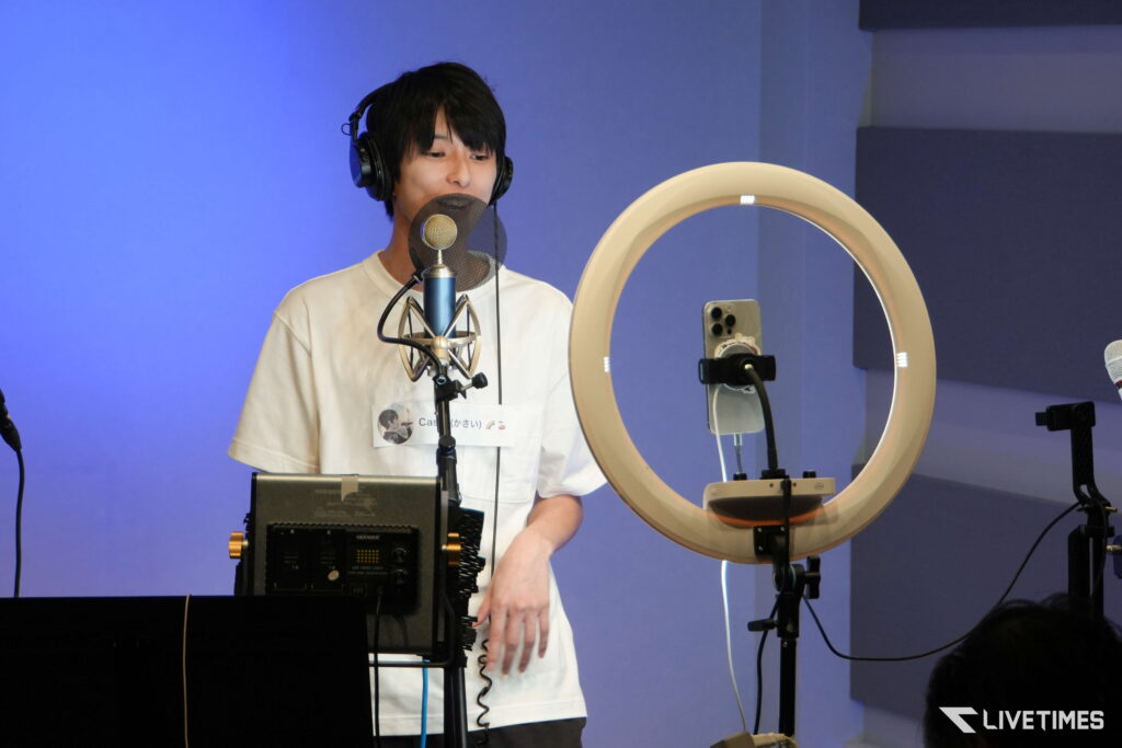 Young man wearing headphones and a white shirt sings into a studio microphone with a pop filter, in a blue recording booth with a circular ring light nearby.