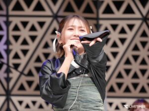 Female singer on stage, mid-performance, holding a microphone and a face shield, wearing a black leather jacket against a geometric backdrop