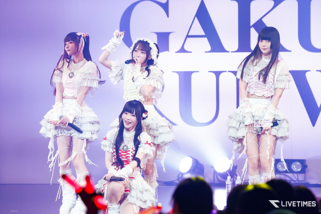 A Japanese idol group performing on stage in frilly white outfits, microphones in hand, with bright purple backdrop and cheering audience in the foreground