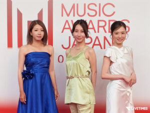 Three women in formal dresses pose on a red carpet background at a Music Awards Japan event in stylish gowns (blue, green, and white).