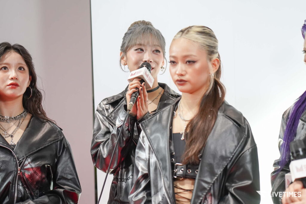 Three women in black leather jackets on stage; center woman speaks into a microphone at a press event.