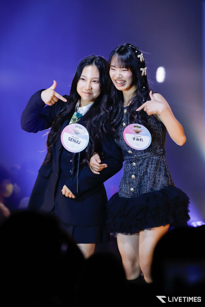 Two young performers on stage smile and point to their round name badges labeled SENJU (left) and すみれ (right). The purple-lit background highlights them.