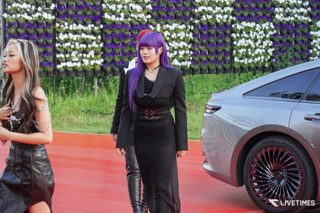 Woman with purple hair in a black blazer and skirt stands beside a silver car, a wall of purple and white flowers behind her.
