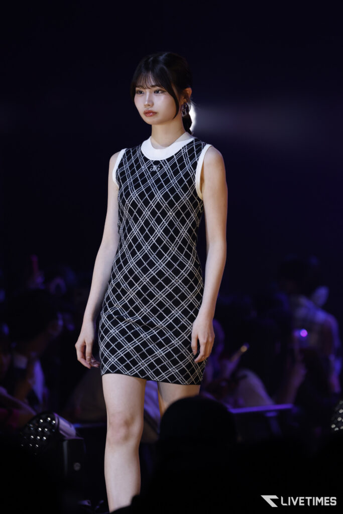 Model walks the runway in a black-and-white plaid sleeveless dress with a white collar, spotlighted on a dark stage.