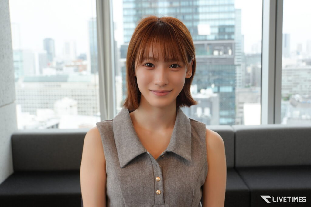 Portrait of a woman with short brown hair wearing a gray sleeveless blouse, seated on a sofa with a city skyline behind her.