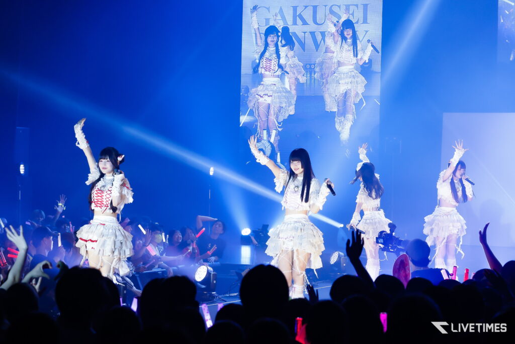 Group of female pop performers in white outfits on a brightly lit stage, raising arms to the audience with blue lighting and a large screen behind them.