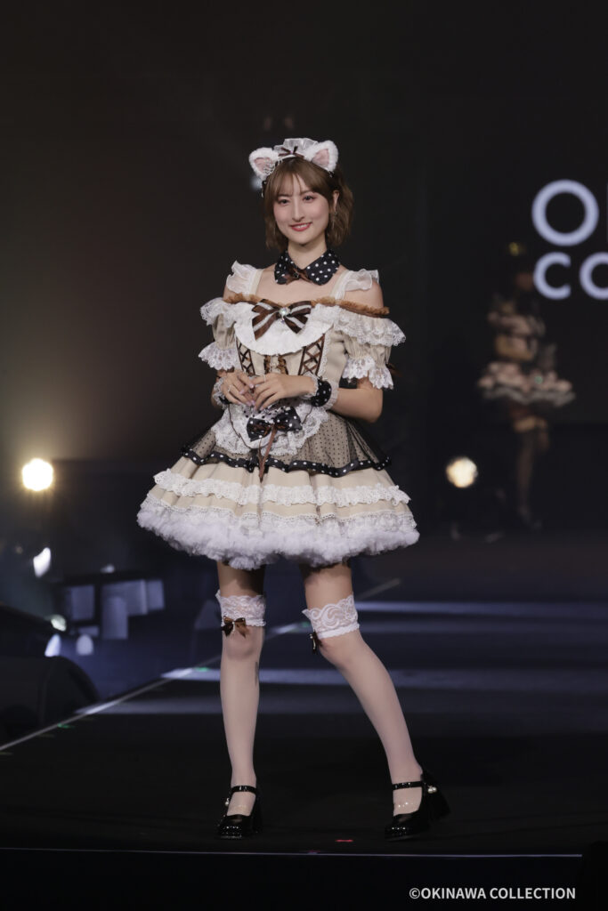 Young woman in a cream-and-brown lace maid costume with cat-ear headband, standing on a dark stage and smiling.