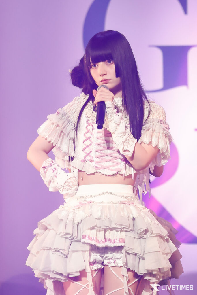 Female performer with long black hair in a white, ruffled dress holding a microphone on a purple-lit stage.