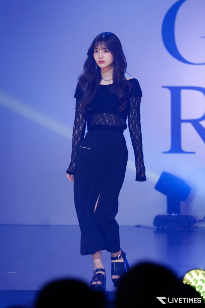 Female performer walking on a blue-lit stage in a black lace top and black pants, with long wavy hair and stage lighting in the background.