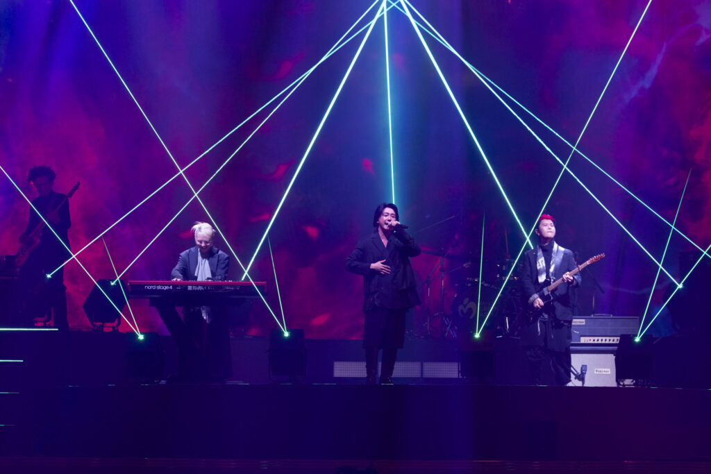 Band performing on stage with neon laser beams crossing the background, center singer with microphone and keyboard player to left, guitarist to right, purple and blue lighting.