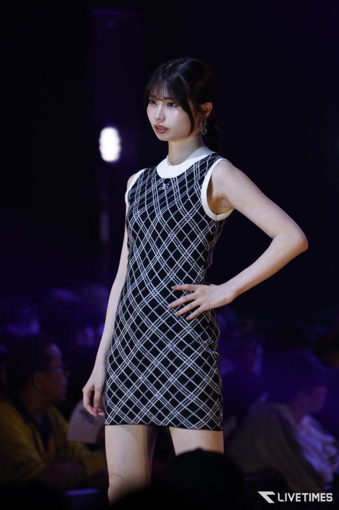 Female fashion model walking the runway in a sleeveless black-and-white plaid dress, with purple stage lighting and an audience in the background