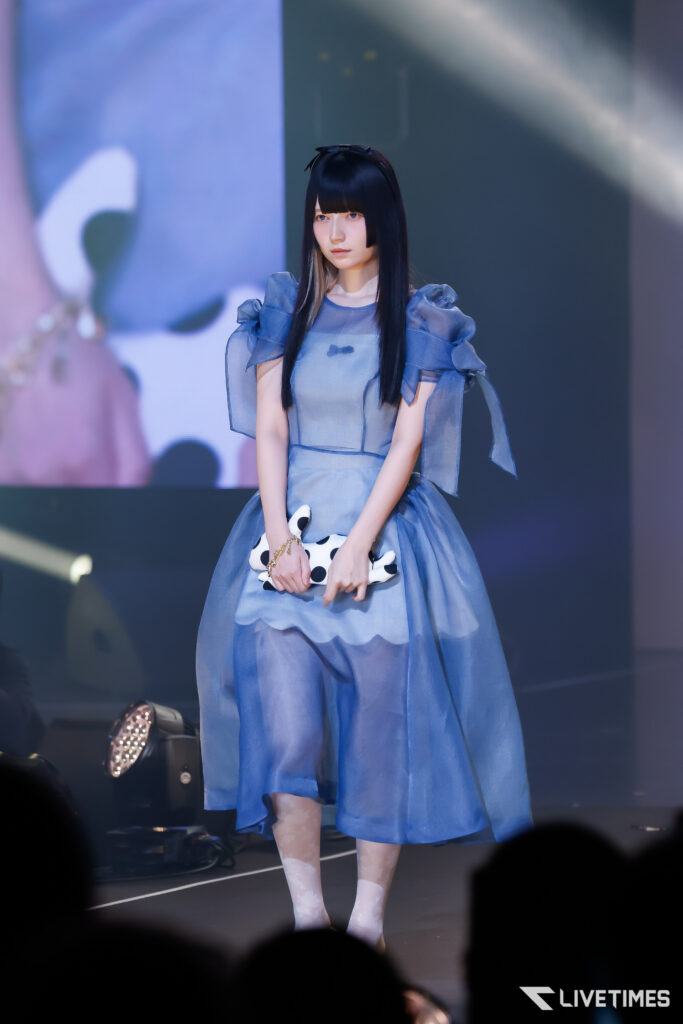 Young woman on stage in a light blue puff-sleeve dress, holding a white polka-dot clutch handily in front of her.