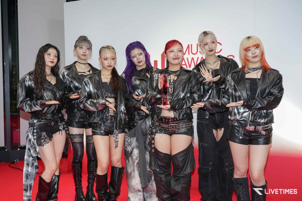 Group of seven women in black leather outfits posing on a red carpet, one holding a glass trophy; backdrop shows red text partials.
