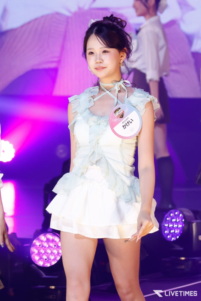 Young woman on stage wearing a light blue frilly dress, with a round name badge pinned to her chest, purple stage lights in the background.