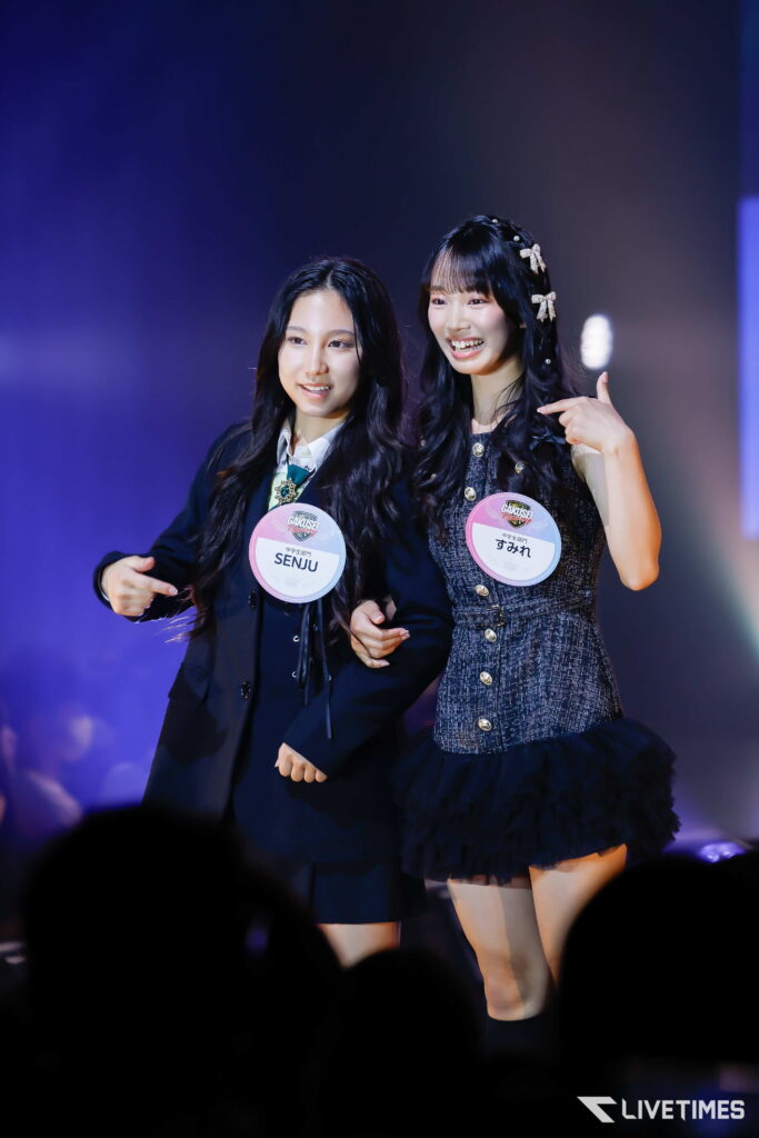 Two young women on stage posing together, each wearing round event badges and smiling under blue-purple stage lights.