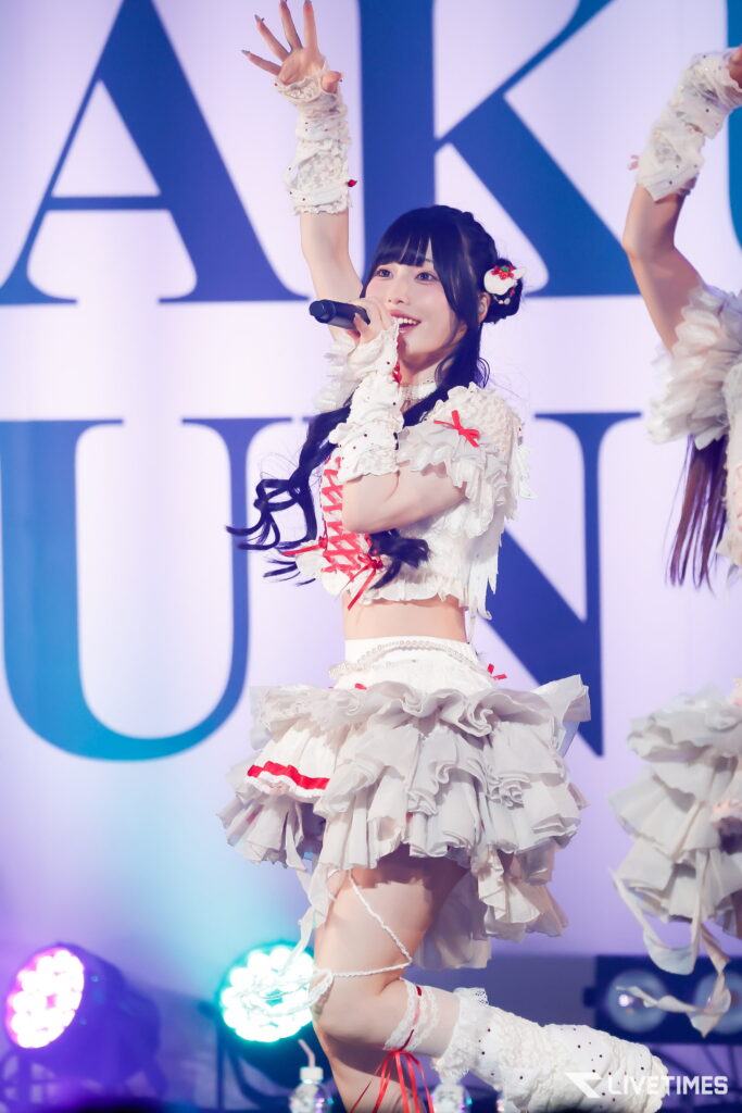 Female performer singing into a microphone with one arm raised, wearing a frilly white costume with red ribbons on stage.