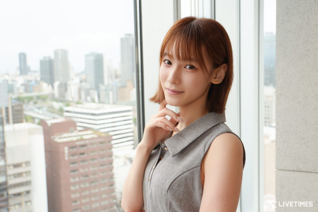 Portrait of a woman with short brown hair in a gray sleeveless top, standing by a window with a city skyline outside the glass.