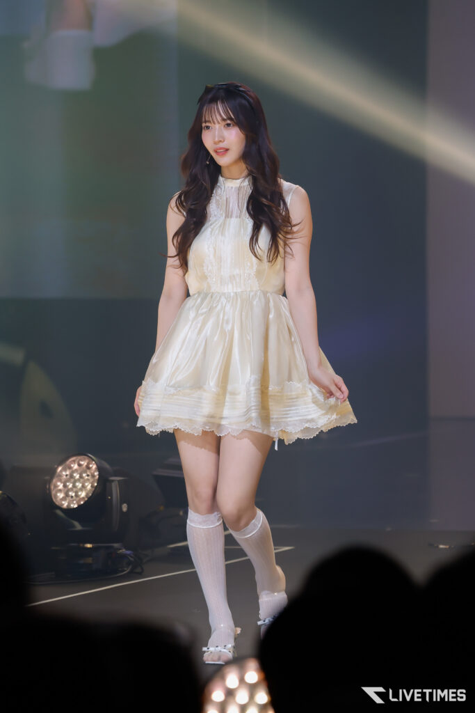 Young woman in a cream lace dress on stage under spotlight during a performance.