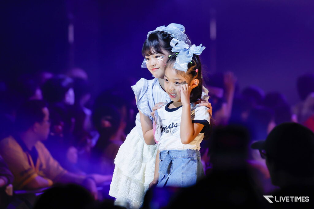 Two young girls pose on stage under purple lights, wearing matching blue hair bows and light outfits while an audience watches.Photo shows a cheerful moment during a performance.