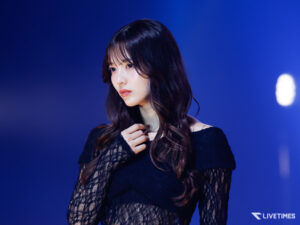 Portrait of a woman with long dark hair in a black lace top, on a blue-lit stage, looking to the left.