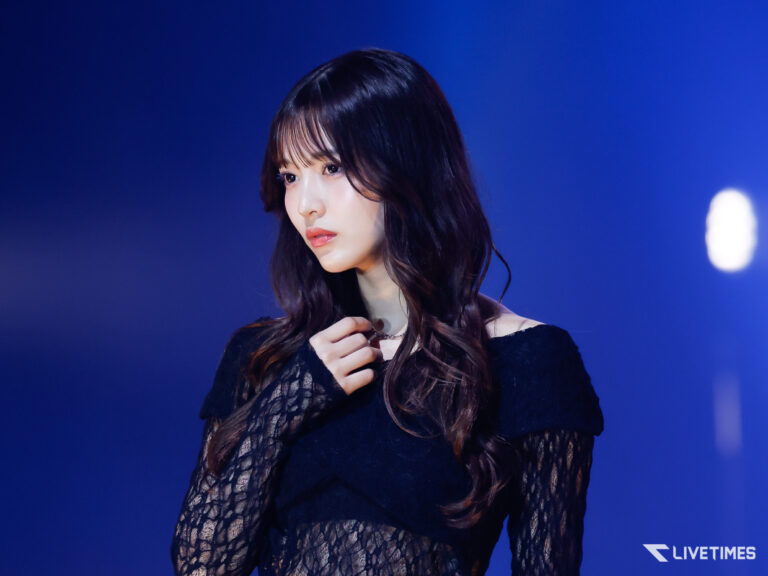 Portrait of a woman with long dark hair in a black lace top, on a blue-lit stage, looking to the left.