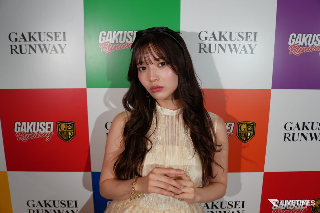 Young woman posing at a colorful backdrop with 'GAKUSEI RUNWAY' logos, wearing a cream lace dress and headband.