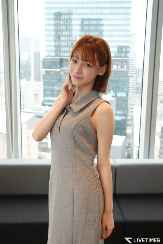 Young woman in a gray sleeveless dress posing by large office windows with a city skyline behind her