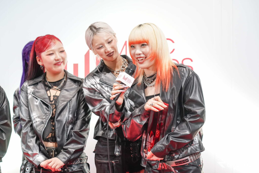 Three women with colorful hair in black leather outfits being interviewed, one holding a microphone with a logo in red text.