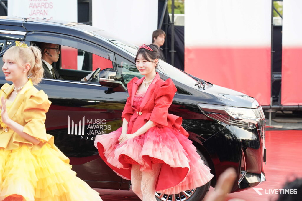 Two women in bright formal gowns pose beside a black SUV at a Music Awards Japan event; pink dress with tiara in foreground, yellow dress partially visible to left, driver inside the car wearing a headset.