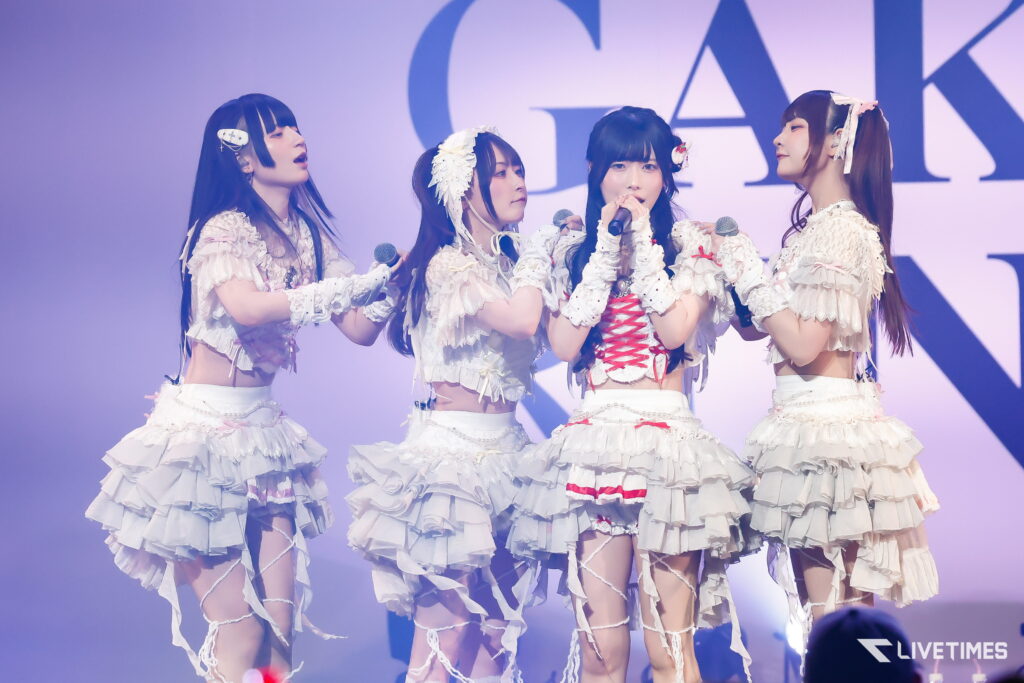 Four female idols in white frilly outfits singing on stage with microphones, under purple stage lighting.