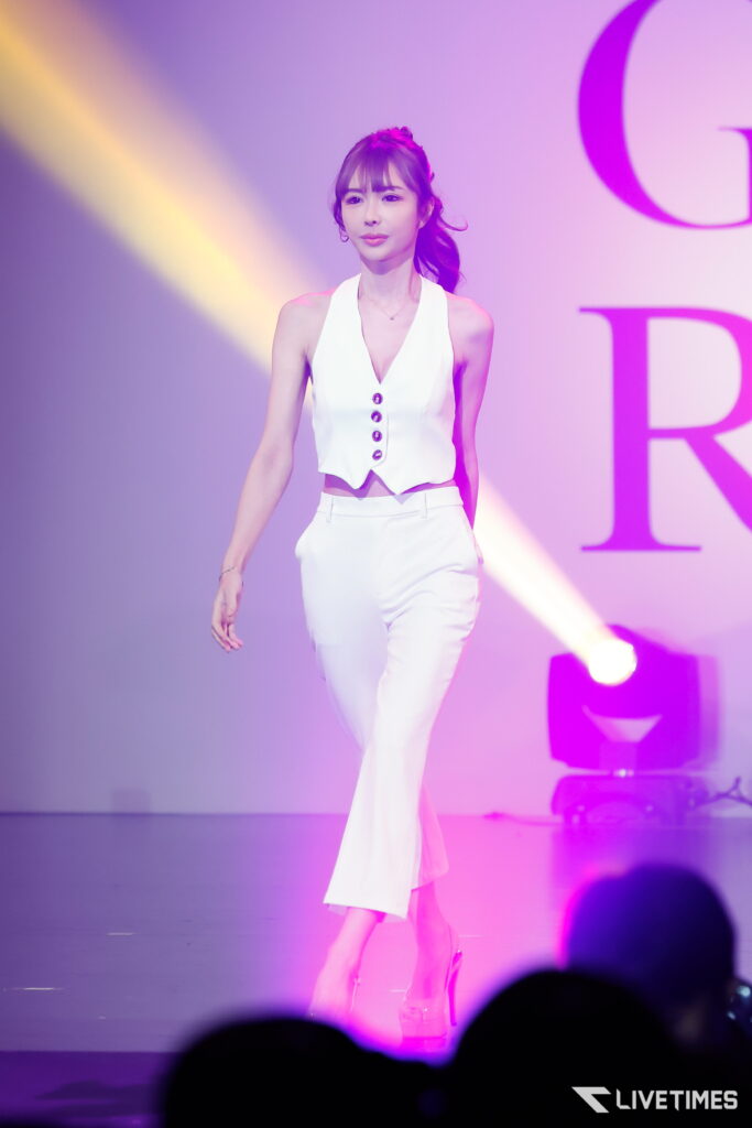 Female performer in a white sleeveless outfit walking on stage under purple/yellow lights, audience silhouettes visible in the foreground.