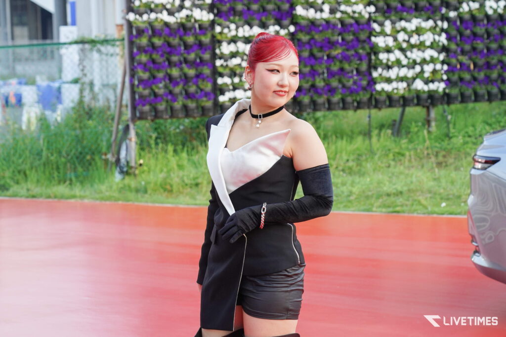 Woman with red hair in a black and white dress posing outdoors with a purple‑white flower wall behind.