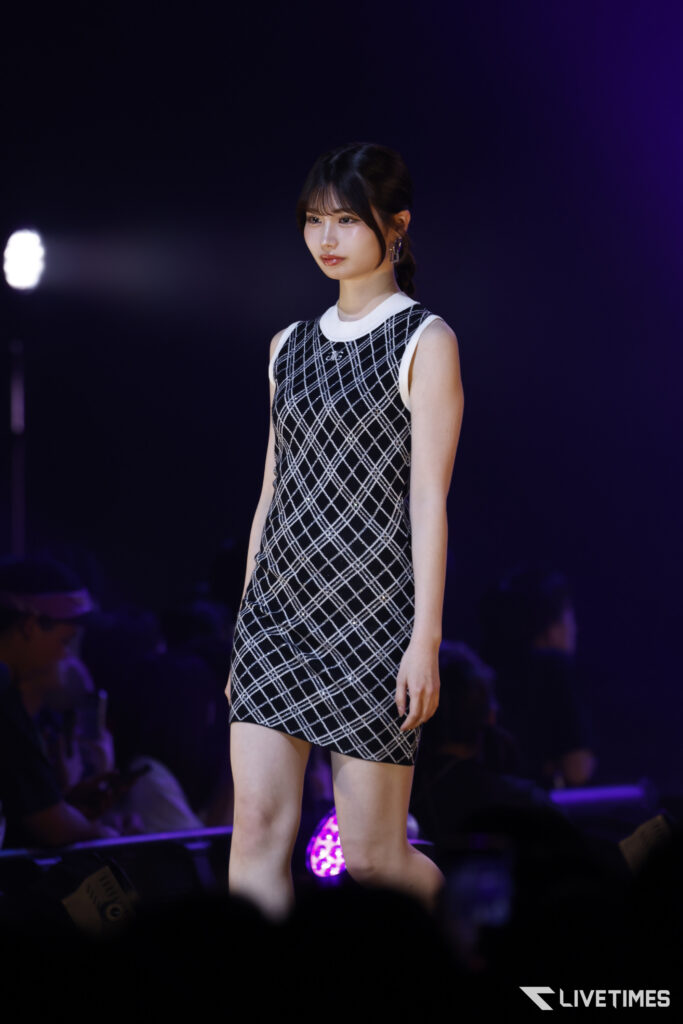 Model walking a fashion-show runway in a black-and-white plaid sleeveless dress under purple stage lights.