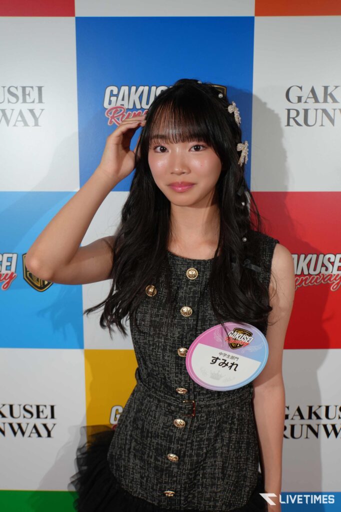 Young woman with long dark hair and bangs posing at a backdrop, wearing a textured black dress with gold buttons and a circular event badge on her chest.