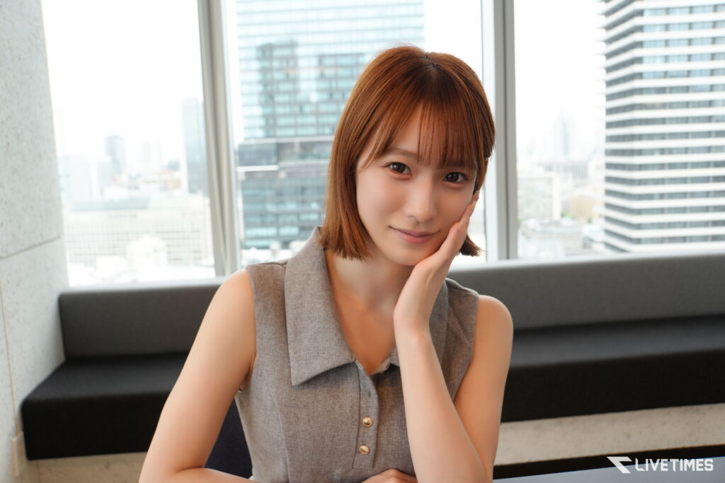 Woman with short brown hair smiling softly, resting her chin on her hand in a bright lounge, city skyscrapers visible through a large window behind her.