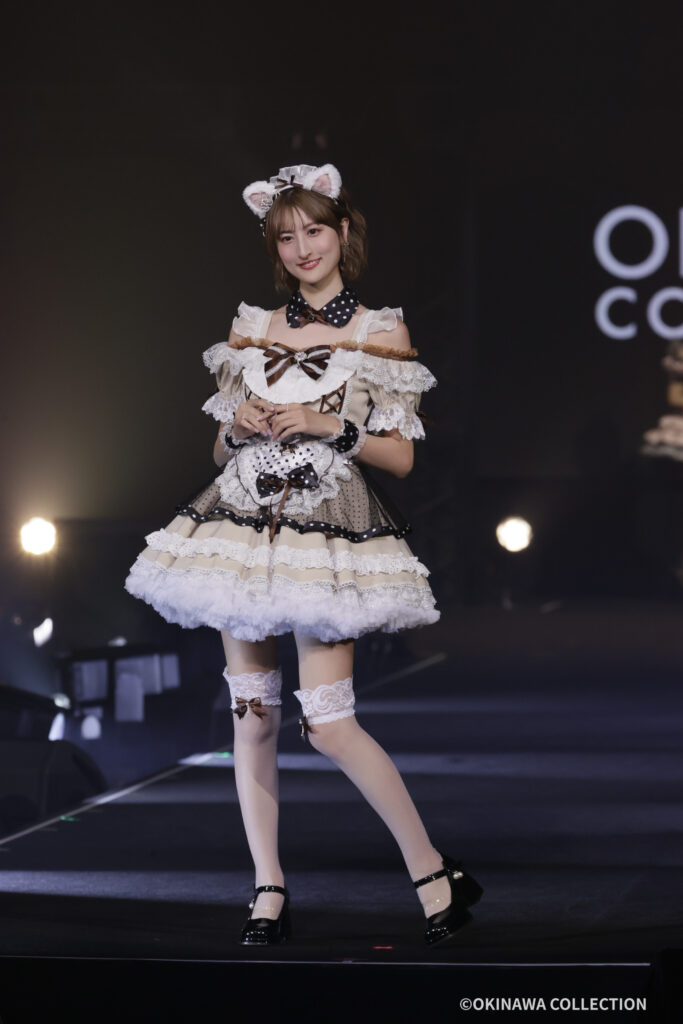 Stage performer in a cream and black lace lolita dress with a cat-ear headband, posing on a dark stage.