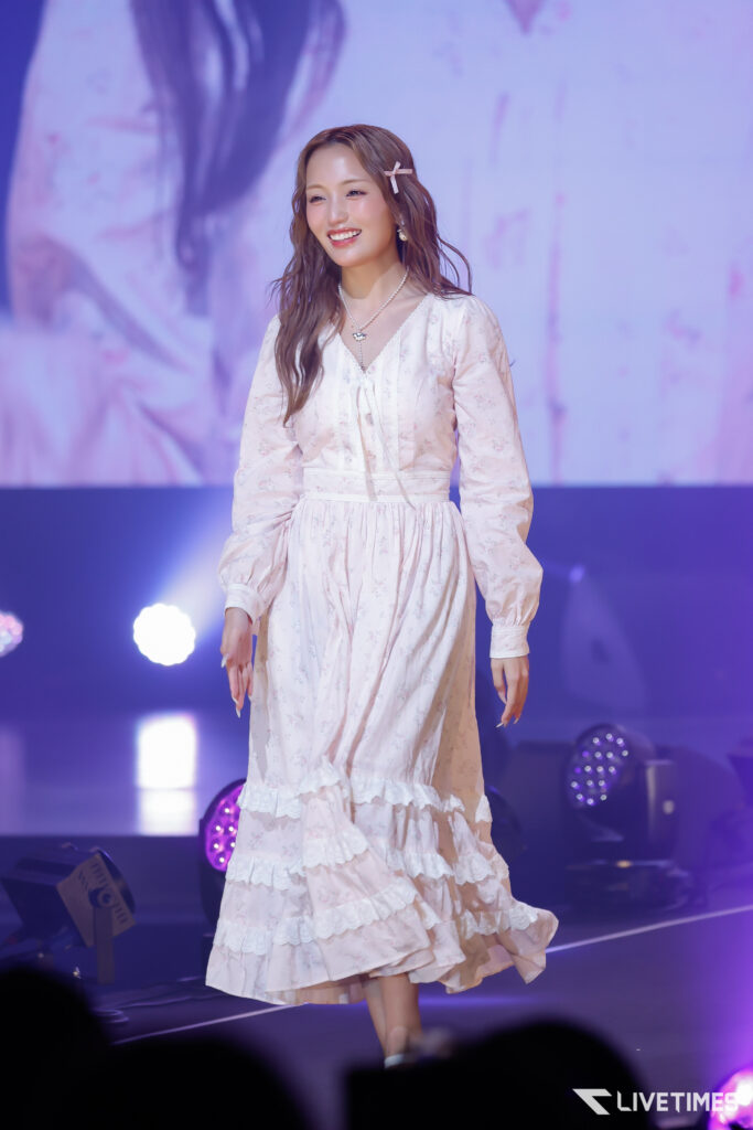 Woman in a light pink floral dress on stage, smiling with hair clipped, under purple stage lighting.