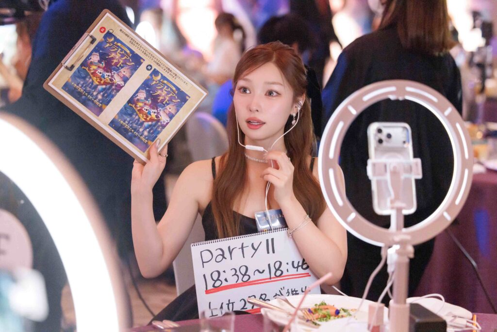 Young woman wearing a headset holds a poster and signs at a booth during a lively event.