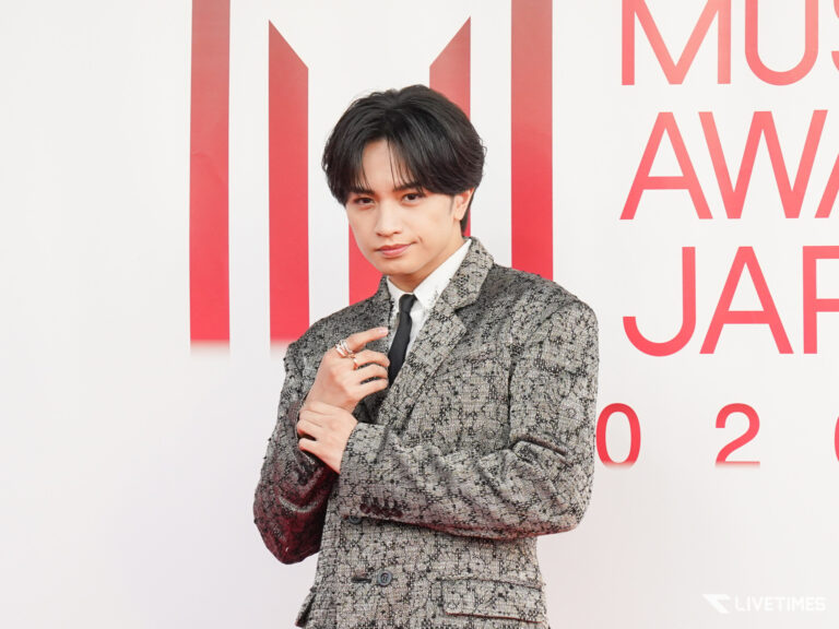 Young man in a metallic-patterned suit posing with hands together against a white backdrop with red text elements behind him at an event.