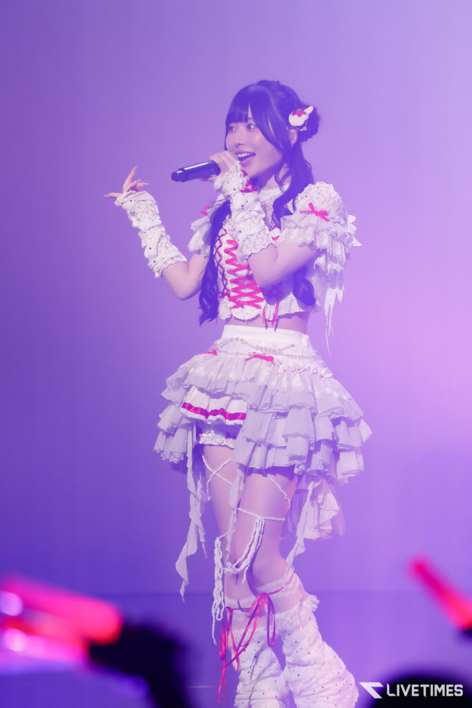 Female singer performs on stage, wearing a white frilly outfit and holding a microphone under purple lighting.