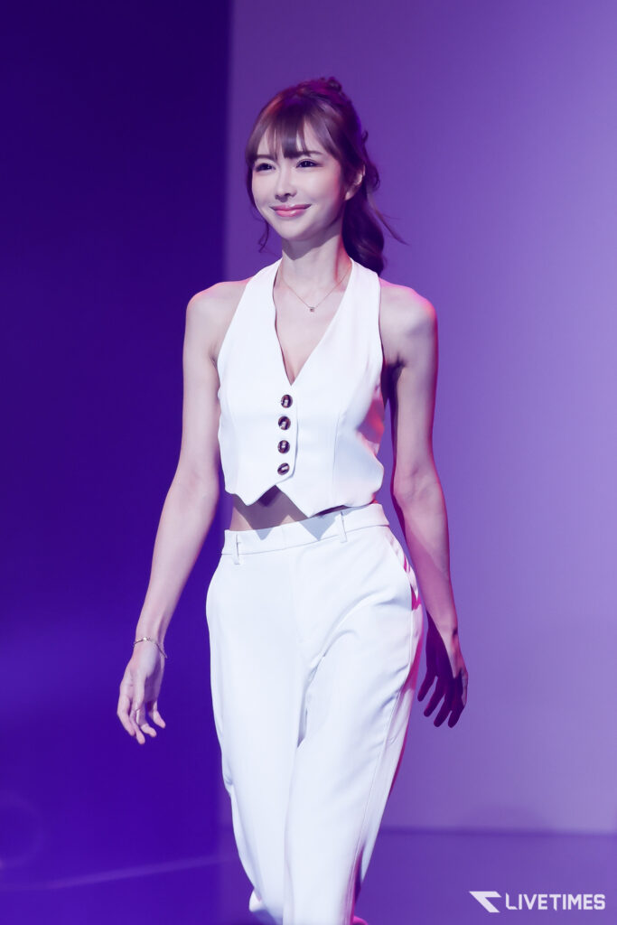 Woman in a white sleeveless top and pants on stage, smiling under purple lighting on stage.