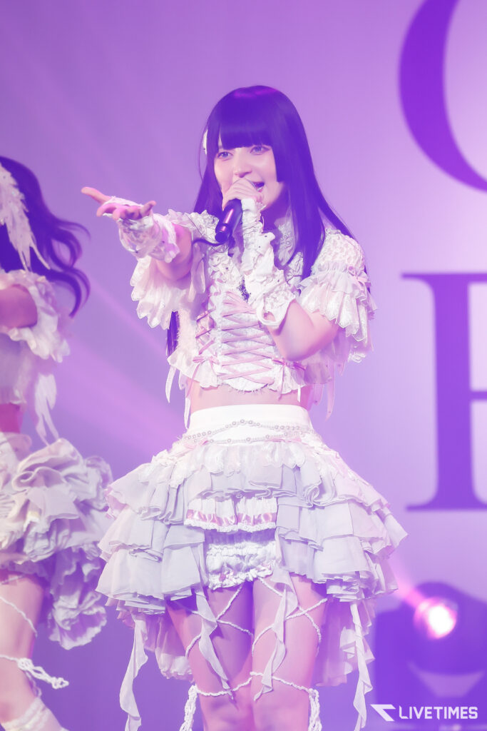 Female performer on stage singing into a microphone, wearing a frilly white costume with ribbons and lace, reaching toward the audience under purple stage lighting.