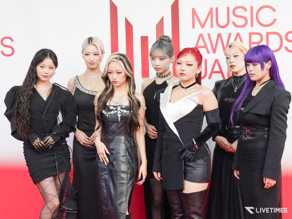 Group of women in black outfits posing on a red carpet at an awards backdrop with bold red text.