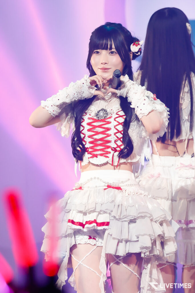 Young female idol on stage making a heart with her hands, wearing a white frilly dress with red corset lacing.