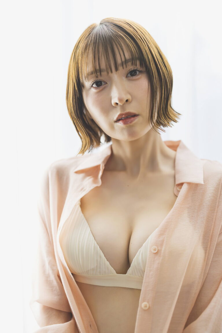 Close-up portrait of a woman with short brown hair and bangs, wearing a pale pink cardigan open over a cream bralette, looking at the camera.