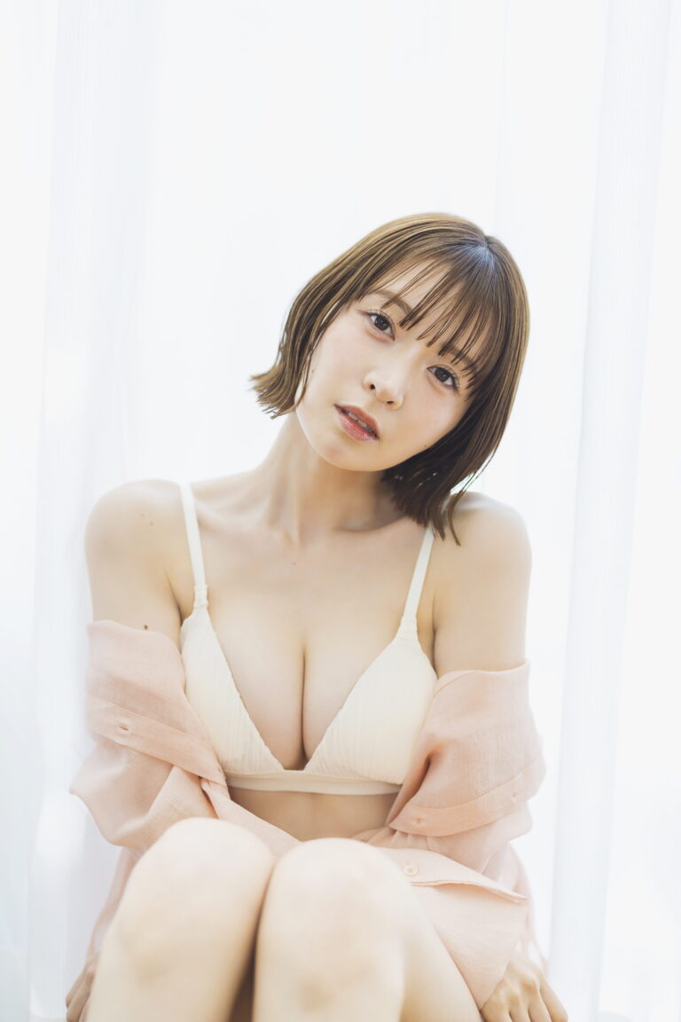 Portrait of a woman with short brown hair in a cream bralette and pale pink cardigan, against white curtains.