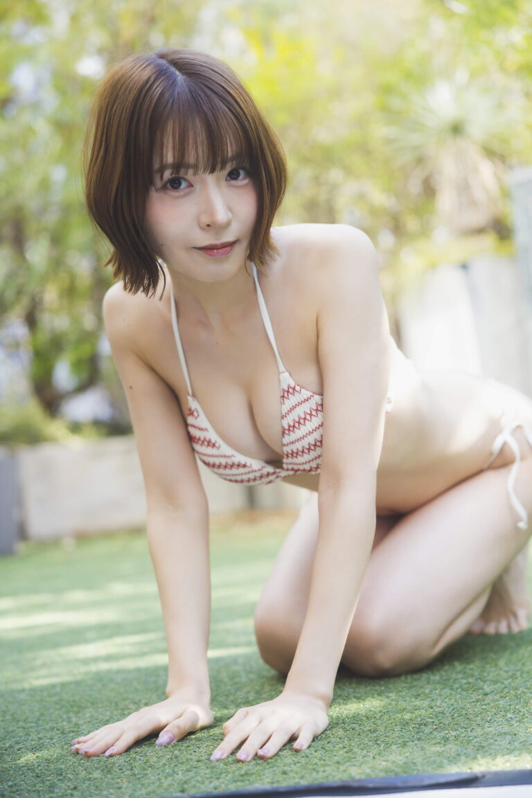 Young woman in a white and red bikini crouching on a grassy outdoor area, hands on the lawn, looking at the camera.