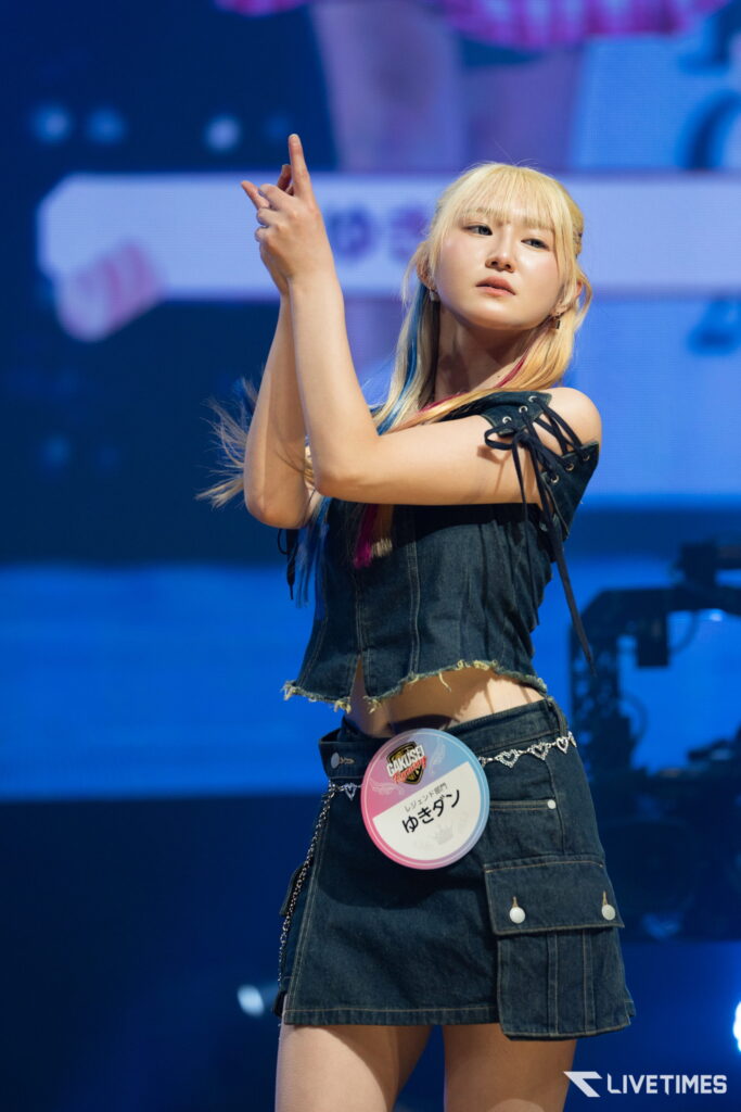 Female performer on stage in a denim outfit, arms raised in a dynamic pose during a concert, badge with text clipped to her waist visible on the right side of her denim skirt.