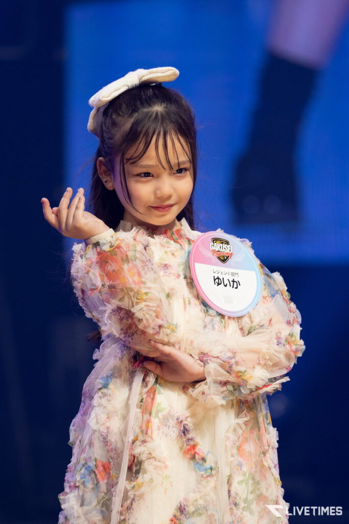 Young girl on stage, wearing a pastel floral dress and a white bow, smiling subtly with a round event badge on her chest.