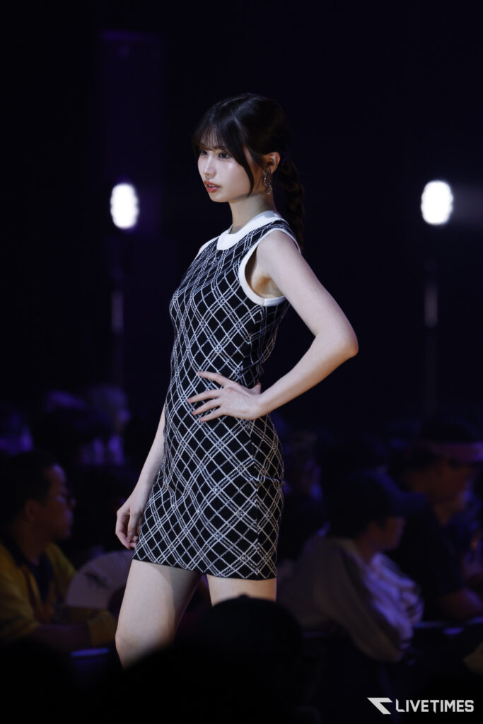 Model walking a runway in a black-and-white plaid sleeveless dress, shown in side profile with hands on hip against a dark stage background.